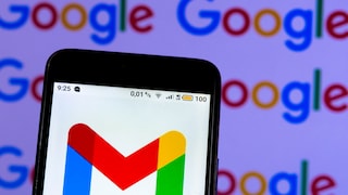 Ein Smartphone wird vor einem unscharfen Hintergrund mit mehrfach dargestelltem Google-Schriftzug gehalten. Auf dem Display ist das mehrfarbige Gmail-Logo auf weißem Hintergrund zu sehen. Am oberen Bildschirmrand sind Statussymbole, die Uhrzeit „9:25“, die Anzeige „4G LTE“ sowie ein Akkustand von „100“ sichtbar. Das Smartphone ist leicht schräg ausgerichtet und nimmt einen großen Teil des Bildes ein. Im Hintergrund wiederholen sich die bunten Google-Logos in Blau, Rot, Gelb und Grün.