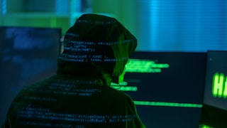 Ein Mann mit Kapuze - vermeintlich ein Hacker - sitzt vor einem Computer und um ihn herum ist ganz viel Datencode.