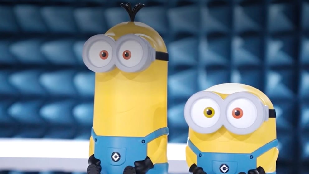 Zwei gelbe Router im Minion-Design stehen nebeneinander auf einer weißen Fläche. Das größere Modell links hat einen längeren Körper, eine graue Brille mit zwei großen Augen, drei schwarze Haarsträhnen auf dem Kopf und eine blaue Latzhose. Das kleinere Modell rechts besitzt ebenfalls eine graue Brille, zwei unterschiedlich gefärbte Augen und eine blaue Latzhose. Beide Geräte haben schwarze Hände und Füße. Im Hintergrund ist eine unscharfe blau gefärbte Struktur mit rechteckigen Formen zu sehen.