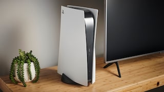 Weiße PlayStation 5 steht senkrecht auf einem Holz-TV-Möbel neben einem Flachbildfernseher. Links befindet sich eine Topfpflanze mit herabhängenden grünen Blättern. Die Spielkonsole zeigt nach vorn, Anschlüsse und Laufwerk sind an der Seite sichtbar. Der Wohnraum ist schlicht eingerichtet und dezent beleuchtet.