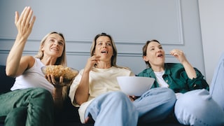 Drei Personen sitzen auf einem Sofa und blicken auf einen Bildschirm außerhalb des Bildes. Eine Person hält eine Schüssel mit Popcorn, zwei weitere halten Snackschüsseln. Die Aufnahme zeigt die Gruppe aus einer niedrigen Perspektive in einem hellen Wohnraum.