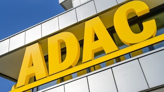 Große gelbe ADAC-Buchstaben an der Fassade eines modernen Gebäudes. Die Aufnahme zeigt das Logo schräg von unten vor blauem Himmel. Die Gebäudefassade besteht aus hellen Metall- und Glaselementen.