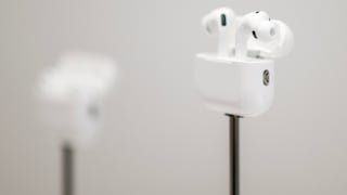 Weiße Apple AirPods Pro in einem geöffneten Ladecase auf einem dünnen schwarzen Ständer. Im Hintergrund ist ein weiteres AirPods-Case unscharf zu sehen. Das Foto ist hell gehalten und zeigt die Kopfhörer vor einem grauen Hintergrund mit geringer Tiefenschärfe.