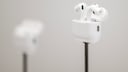 Wei&szlig;e Apple AirPods Pro in einem ge&ouml;ffneten Ladecase auf einem d&uuml;nnen schwarzen St&auml;nder. Im Hintergrund ist ein weiteres AirPods-Case unscharf zu sehen. Das Foto ist hell gehalten und zeigt die Kopfh&ouml;rer vor einem grauen Hintergrund mit geringer Tiefensch&auml;rfe.