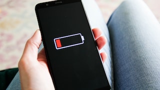 Eine Person hält ein Smartphone in der Hand. Auf dem schwarzen Display ist ein nahezu leerer Akku mit roter Ladeanzeige zu sehen. Im Hintergrund sind unscharf die Beine der Person erkennbar.
