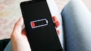 Eine Person h&auml;lt ein Smartphone in der Hand. Auf dem schwarzen Display ist ein nahezu leerer Akku mit roter Ladeanzeige zu sehen. Im Hintergrund sind unscharf die Beine der Person erkennbar.