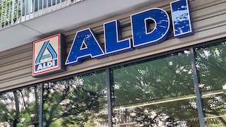Außenansicht einer Aldi-Filiale mit großem blauem Aldi-Schriftzug und Aldi-Logo an der Gebäudefassade. Unter dem Schild befinden sich große Fenster, in denen sich Bäume und ihre Blätter spiegeln. Oberhalb des Eingangs ist ein Balkon mit Metallgeländer zu sehen.