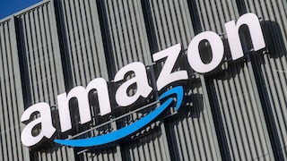 Schräg aufgenommene Ansicht eines großen Amazon-Logos mit blauem Pfeil an einer Fassade aus vertikalen Metallpaneelen vor blauem Himmel.