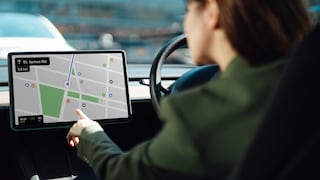 Person sitzt am Steuer eines Autos und bedient einen großen Bildschirm auf dem Armaturenbrett. Auf dem Display ist eine Kartenansicht einer Navigations-App mit Route und Straßeninformationen zu sehen. Das Lenkrad befindet sich rechts neben dem Bildschirm, im Hintergrund ist unscharfer Verkehr erkennbar.