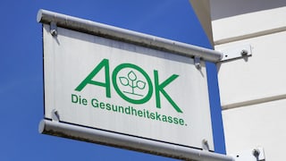 AOK-Schild mit grünem Logo und Schriftzug „Die Gesundheitskasse“ an einer weißen Gebäudefassade vor blauem Himmel.