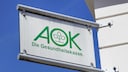 AOK-Schild mit gr&uuml;nem Logo und Schriftzug &bdquo;Die Gesundheitskasse&ldquo; an einer wei&szlig;en Geb&auml;udefassade vor blauem Himmel.