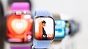 Eine Apple Watch mit hellblauem Armband steht im Vordergrund. Auf dem Display ist ein Zifferblatt mit einer Person im Profil zu sehen. Im unscharfen Hintergrund befinden sich weitere Apple Watches mit unterschiedlichen Armb&auml;ndern und farbigen Anzeigen.