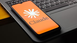 Ein Smartphone mit orangefarbenem Claude-Startbildschirm liegt schräg auf der Tastatur eines aufgeklappten Laptops. Auf dem Display sind das Claude-Logo und der Schriftzug „Claude“ zu sehen. Die Aufnahme zeigt das Gerät in Nahansicht auf einem dunklen Tisch.