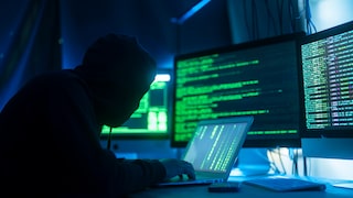 Person mit Kapuzenpullover sitzt vor mehreren Monitoren und einem Laptop. Auf den Bildschirmen sind grün dargestellte Programmzeilen und Systeminformationen zu sehen. Die Szene ist in blaues und grünes Licht getaucht und zeigt einen Arbeitsplatz in einer IT-Umgebung.