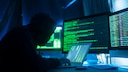 Person mit Kapuzenpullover sitzt vor mehreren Monitoren und einem Laptop. Auf den Bildschirmen sind gr&uuml;n dargestellte Programmzeilen und Systeminformationen zu sehen. Die Szene ist in blaues und gr&uuml;nes Licht getaucht und zeigt einen Arbeitsplatz in einer IT-Umgebung.