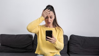 Person sitzt auf einem Sofa und blickt auf ein Smartphone in der Hand, während eine Hand auf der Stirn liegt. Die Person trägt einen gelben Pullover und eine Jeans. Im Hintergrund ist eine helle Wand zu sehen.