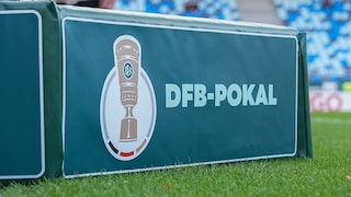 DFB-Pokal-Logo auf einer grünen Werbebande am Spielfeldrand eines Fußballstadions. Im Hintergrund sind unscharfe blaue und weiße Tribünensitze sowie Teile des Rasens zu sehen.