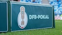 DFB-Pokal-Logo auf einer gr&uuml;nen Werbebande am Spielfeldrand eines Fu&szlig;ballstadions. Im Hintergrund sind unscharfe blaue und wei&szlig;e Trib&uuml;nensitze sowie Teile des Rasens zu sehen.
