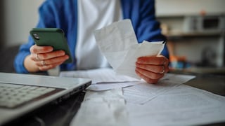 Eine Person sitzt an einem Tisch und hält ein Smartphone sowie mehrere Papierbelege in den Händen. Auf dem Tisch liegen weitere Dokumente und links im Vordergrund ist ein aufgeklappter Laptop zu sehen. Der Hintergrund ist unscharf.