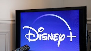 Fernseher mit Disney+-Logo auf blauem Hintergrund, davor eine Hand mit Fernbedienung vor dem Bildschirm.