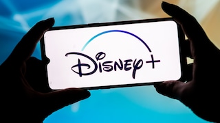 Zwei Hände halten ein Smartphone im Querformat. Auf dem hellen Display ist groß das Disney+-Logo zu sehen. Im unscharfen Hintergrund dominieren Blau-, Türkis- und helle Beigetöne.