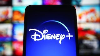 Ein Bildschirm mit dem Disney+-Logo steht im Vordergrund. Im Hintergrund sind unscharf mehrere bunte Vorschaubilder von Filmen und Serien auf einer Streaming-Oberfläche zu sehen.