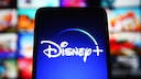 Ein Bildschirm mit dem Disney+-Logo steht im Vordergrund. Im Hintergrund sind unscharf mehrere bunte Vorschaubilder von Filmen und Serien auf einer Streaming-Oberfl&auml;che zu sehen.