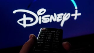 Eine Fernbedienung wird vor einen Fernseher gehalten. Auf dem Bildschirm ist unscharf das Disney+-Logo auf blauem Hintergrund zu sehen.