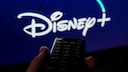 Eine Fernbedienung wird vor einen Fernseher gehalten. Auf dem Bildschirm ist unscharf das Disney+-Logo auf blauem Hintergrund zu sehen.