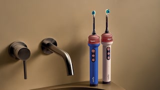 Zwei elektrische Zahnbürsten von Dyson stehen auf dem Rand eines Waschbeckens neben einem wandmontierten Wasserhahn. Eine Zahnbürste ist blau, die andere hellrosa. Beide besitzen im oberen Bereich einen transparenten Abschnitt unter dem Bürstenkopf. Im Hintergrund ist eine beige Wand zu sehen.