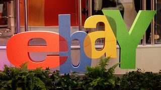 Großes farbiges Ebay-Logo mit roten, blauen, gelben und grünen Buchstaben vor einem Messestand. Im Vordergrund stehen mehrere Pflanzen, im Hintergrund sind Glaswände und Teile der Ausstellungsfläche zu sehen.