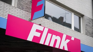 Das Bild zeigt den Eingangsbereich eines Flink-Standorts. Auffällig sind das große pinkfarbene Flink-Logo und der gleichfarbige Schriftzug an der Fassade. Darüber sind große Fenster sowie eine Fassade aus weißen und grauen Backsteinen zu sehen.