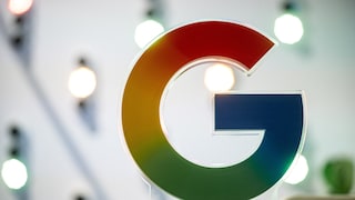 Großes Google-Logo in den Unternehmensfarben Rot, Gelb, Grün und Blau. Das Logo steht auf einer transparenten Halterung im Vordergrund. Im unscharfen Hintergrund sind mehrere helle Lichtquellen in einem Innenraum zu sehen.