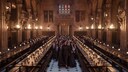 Gro&szlig;e Halle von Hogwarts mit langen Holztischen und zahlreichen Sch&uuml;lerinnen und Sch&uuml;lern in Schuluniformen. Im Mittelgang gehen mehrere Personen zwischen den Tischreihen hindurch. Fackeln beleuchten den hohen Saal mit Bannern und Fenstern im Hintergrund.