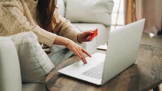 Eine Person sitzt mit einem Laptop an einem Holztisch und hält eine rote Karte in der Hand. Auf dem Tisch liegt außerdem ein Smartphone. Im Hintergrund sind ein helles Sofa und ein Fenster zu sehen.