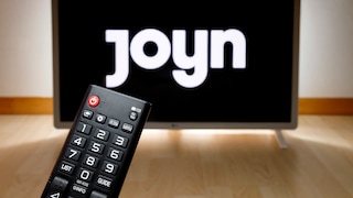 Eine Fernbedienung liegt vor einem Fernseher. Auf dem Bildschirm im Hintergrund ist das Logo des Streaming-Dienstes Joyn zu sehen.