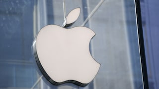 Nahaufnahme eines weißen Apple-Logos an einer Glaswand. Im Hintergrund sind verschwommene Strukturen eines modernen Gebäudes in Blau- und Grautönen zu sehen.