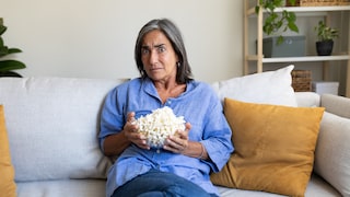 Person in lockerem blauem Hemd sitzt auf einem hellen Sofa und hält eine Glasschüssel mit Popcorn in den Händen. Im Hintergrund stehen ein Regal mit Pflanzen, Aufbewahrungsboxen und Dekoration. Auf dem Sofa liegen mehrere beige und senffarbene Kissen. Die Person blickt direkt in die Kamera. Die Aufnahme entstand in einem hellen Wohnzimmer.