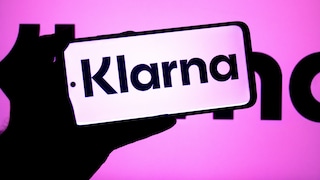 Eine Person hält ein Smartphone mit dem Klarna-Logo vor einen pink-violetten Hintergrund, auf dem ebenfalls das Klarna-Logo zu sehen ist. Das Display zeigt den Schriftzug „Klarna“ in schwarzer Schrift auf hellem Hintergrund.
