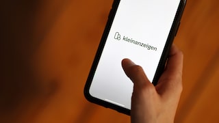 Eine Hand hält ein Smartphone, auf dessen Bildschirm das grüne Logo von Kleinanzeigen zu sehen ist. Der Hintergrund ist in Orange- und Brauntönen gehalten.