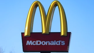 Das Bild zeigt ein großes McDonald’s-Schild vor einem wolkenlosen blauen Himmel. Im oberen Bereich sind die gelben, bogenförmigen Elemente des Logos zu sehen. Darunter befindet sich eine dunkelrote, rechteckige Tafel mit dem weißen Schriftzug „McDonald’s“. Am unteren Bildrand ist ein Teil der schwarzen Halterung des Schildes erkennbar, links außerdem die Spitzen eines kahlen Baumes.