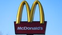 Das Bild zeigt ein gro&szlig;es McDonald&rsquo;s-Schild vor einem wolkenlosen blauen Himmel. Im oberen Bereich sind die gelben, bogenf&ouml;rmigen Elemente des Logos zu sehen. Darunter befindet sich eine dunkelrote, rechteckige Tafel mit dem wei&szlig;en Schriftzug &bdquo;McDonald&rsquo;s&ldquo;. Am unteren Bildrand ist ein Teil der schwarzen Halterung des Schildes erkennbar, links au&szlig;erdem die Spitzen eines kahlen Baumes.