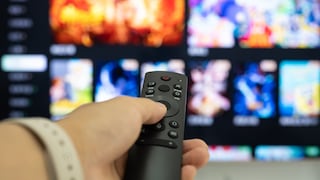 Eine Hand hält eine schwarze Fernbedienung vor einem Fernseher mit einer unscharf dargestellten Streaming-Oberfläche und mehreren Video-Kacheln.
