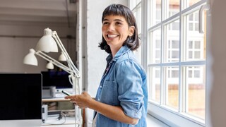 Person in hellem Jeanshemd steht in einem Büro am Fenster und hält ein Smartphone in der Hand. Auf einem Schreibtisch stehen mehrere Monitore, Schreibtischlampen und ein Notizbuch. Der Raum ist hell und wird von großen Fenstern mit Tageslicht beleuchtet.