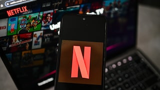 Smartphone mit Netflix-Logo vor einem Laptop. Auf dem Bildschirm des Laptops ist die Netflix-Startseite mit verschiedenen Film- und Serienkacheln zu sehen.