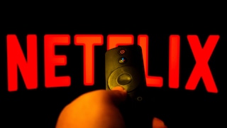 Eine Hand hält eine Fernbedienung vor einem Bildschirm mit groß eingeblendetem roten Netflix-Logo auf schwarzem Hintergrund.