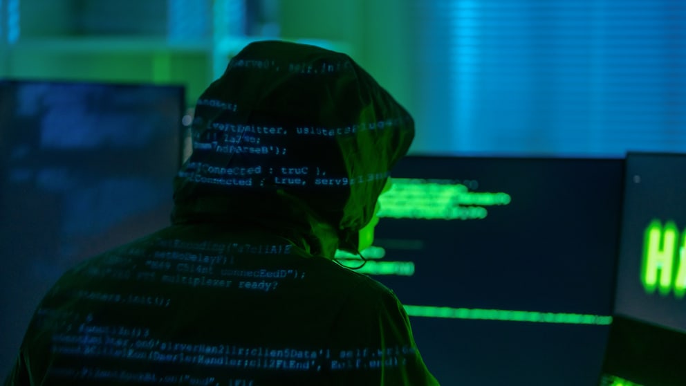 Eine Person mit Kapuze sitzt vor mehreren Computerbildschirmen in einem dunkel beleuchteten Raum. Auf Kleidung und Monitoren sind unscharfe Programmcode-Zeilen zu sehen. Die Szene ist in grünem und blauem Licht gehalten.