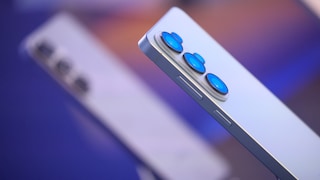 Nahaufnahme der Rückseite eines hellen Samsung-Smartphones mit drei Kameralinsen, im Hintergrund ein unscharfes zweites Gerät derselben Serie.