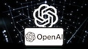 Auf einem Laptop-Bildschirm ist das OpenAI-Logo mehrfach zu sehen. Im Hintergrund verlaufen leuchtende Linien und Punkte zu einem digitalen Netzwerk auf schwarzem Grund. Im Vordergrund erscheint ein wei&szlig;es Feld mit dem OpenAI-Schriftzug und Logo. Die Tastatur des Laptops ist am unteren Bildrand sichtbar.