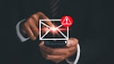 Person h&auml;lt ein Smartphone in der Hand und tippt auf ein eingeblendetes Briefumschlag-Symbol. Daneben ist ein rotes Warnsymbol mit Ausrufezeichen zu sehen. Das Bild visualisiert eine verd&auml;chtige oder potenziell gef&auml;hrliche E-Mail.